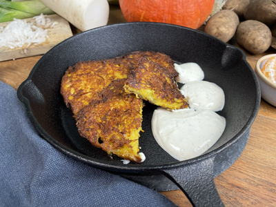 KI generiert: Das Bild zeigt drei goldbraune Reibekuchen in einer schwarzen Pfanne, begleitet von einem Klecks weißem Dip. Im Hintergrund sind verschiedene frische Zutaten wie ein Kürbis, Kartoffeln und ein Rettich zu sehen.