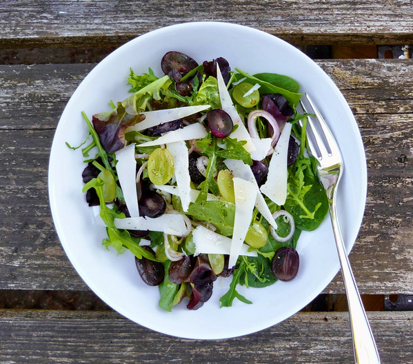 KI generiert: Ein frischer Salat mit Trauben, Käse und grünem Blattgemüse in einer weißen Schale auf Holzuntergrund.