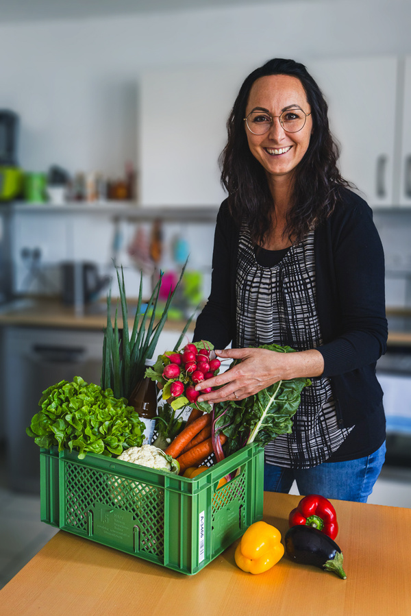 KI generiert: Eine Person steht in einem Gemüsefeld und hält eine Kiste mit frischem Gemüse. Im Hintergrund ist ein Bauernhof zu sehen.