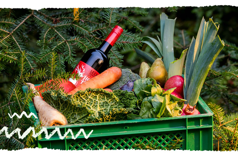 KI generiert: Kiste mit Gemüse, Obst und einer Flasche Wein vor Tannenzweigen.