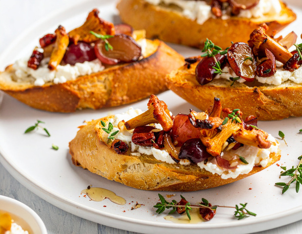 KI generiert: Geröstetes Brot mit Ziegenkäse, Pfifferlingen, Trauben und Kräutern auf einem weißen Teller.