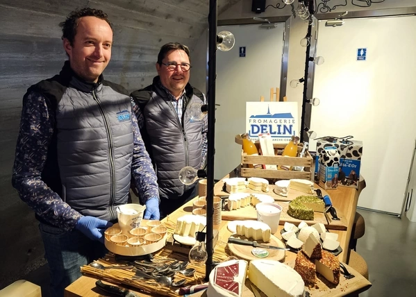 KI generiert: Zwei Männer präsentieren Käsesorten an einem Tisch. Schild: "FROMAGERIE DELIN".