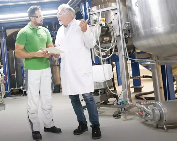 KI generiert: Zwei Männer in einem industriellen Umfeld diskutieren vor Maschinen.