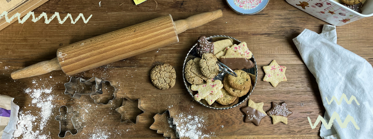 Das Bild zeigt eine Sammlung von Weihnachtsplätzchen auf einem Holztisch, umgeben von Ausstechformen und einem Nudelholz. Ein weißes Tuch und eine Dose ergänzen die Szene.