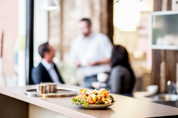 KI generiert: Das Bild zeigt einen Teller mit frischen Obstspießen auf einer Theke im Vordergrund, während unscharf im Hintergrund zwei Personen in einem Gespräch an einer Theke stehen. Der Fokus liegt auf dem leckeren Obstarrangement.