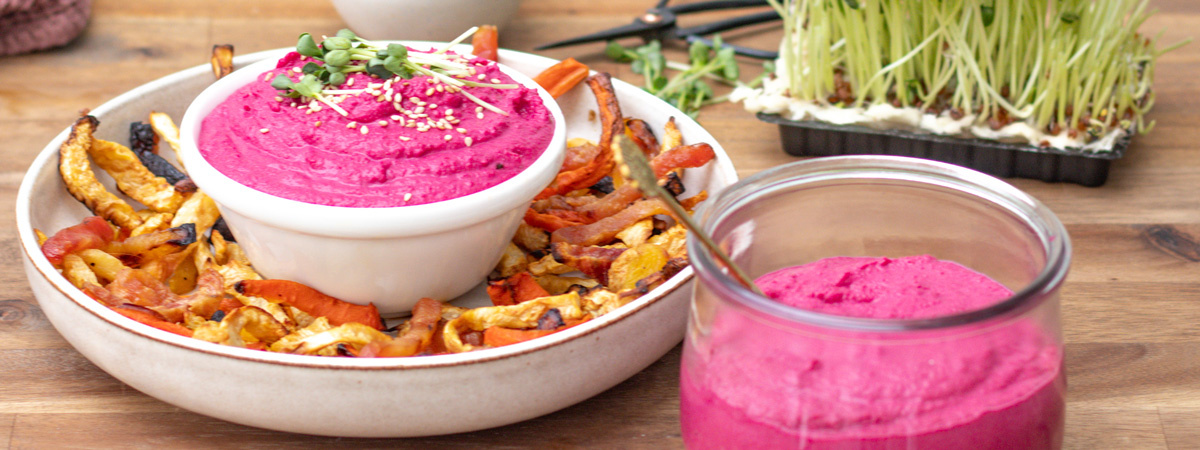 Das Bild zeigt einen Burger aus pflanzlichen Zutaten auf einem weißen Teller, umgeben von weiteren Schüsseln mit grünen Beilagen und einer kleinen Schüssel mit gelber Soße. Der Fokus liegt auf der Präsentation des Burgers mit einem Sesambrötchen, einem grünen Patty und Gemüse.