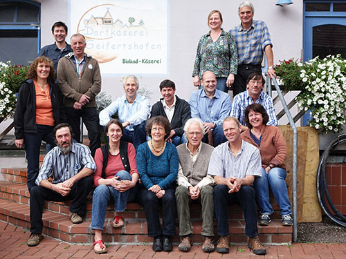 KI generiert: Eine Gruppe von Menschen posiert vor einem Schild: "Dorfsennerei Geifertshofen - Bioland-Käserei".