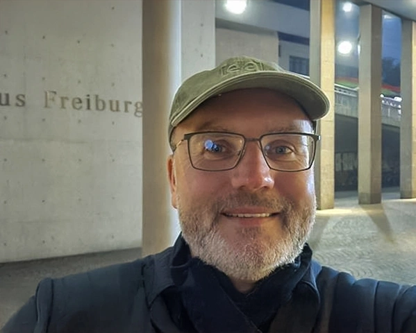 KI generiert: Ein Mann mit Brille und Mütze lächelt vor einem Gebäude. Text: "us Freiburg".