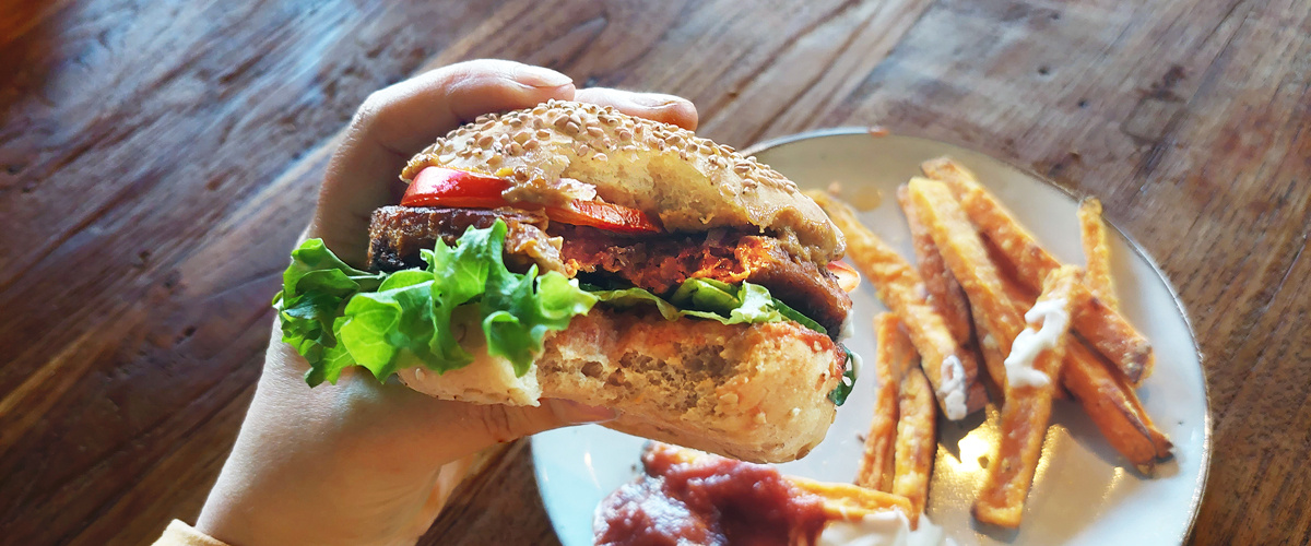 KI generiert: Ein handgehaltener Burger mit Salat, Tomate und Soße; daneben Süßkartoffelpommes.
