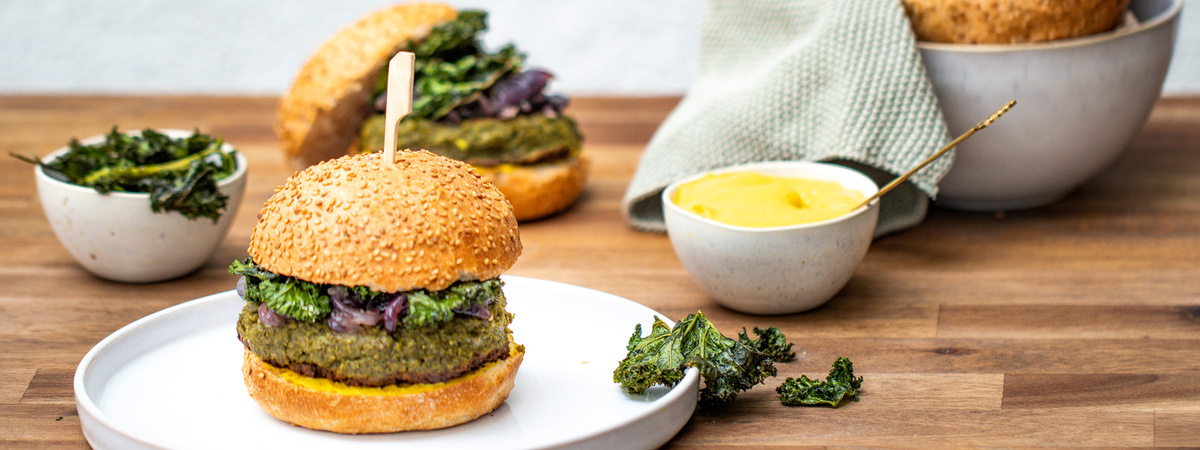 Das Bild zeigt einen Burger aus pflanzlichen Zutaten auf einem weißen Teller, umgeben von weiteren Schüsseln mit grünen Beilagen und einer kleinen Schüssel mit gelber Soße. Der Fokus liegt auf der Präsentation des Burgers mit einem Sesambrötchen, einem grünen Patty und Gemüse.