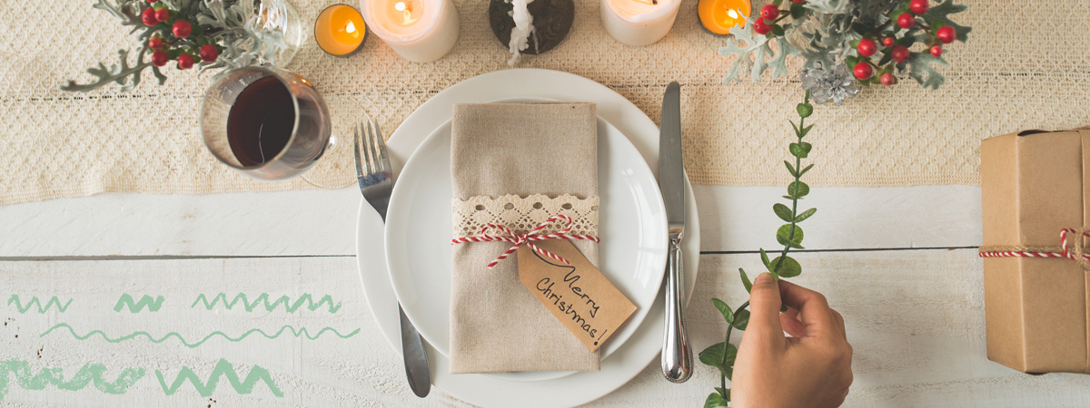 KI generiert: Das Bild zeigt einen festlich gedeckten Tisch mit einem weißen Teller, auf dem eine beige Serviette mit einem "Merry Christmas"-Etikett liegt. Daneben befinden sich Kerzen, ein Glas Rotwein, eine eingepackte Geschenkbox und weihnachtliche Dekorationen.