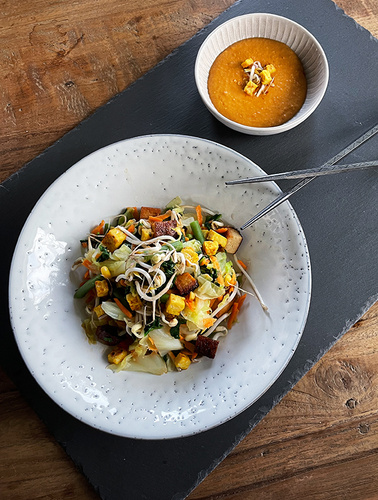 KI generiert: Ein Teller mit einem bunten Gemüsesalat und eine Schüssel mit Suppe auf einem Tisch.