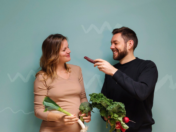KI generiert: Zwei Menschen stehen nebeneinander und halten frisches Gemüse in den Händen, während sie sich gegenseitig freundlich anschauen. Der Hintergrund ist einfarbig mit einem schlichten grafischen Muster.