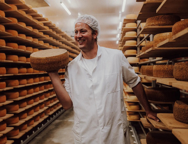 KI generiert: Ein Mann in weißer Kleidung steht in einer Käselagerung und hält einen Käselaib.