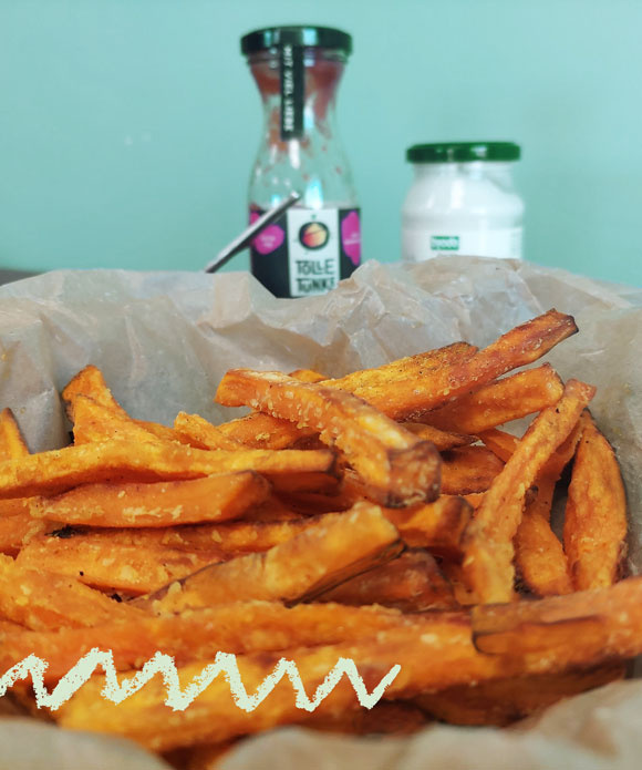 KI generiert: Süßkartoffelpommes in einer Schale mit zwei Soßengläsern im Hintergrund. Text: "Tolle Knolle".