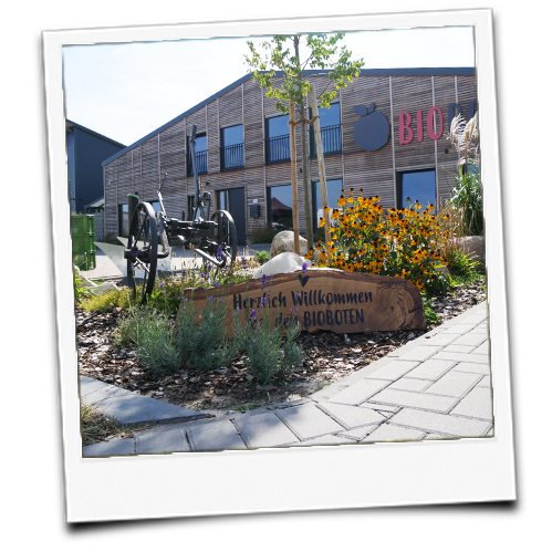KI generiert: Holzgebäude mit Blumenbeet, Schild: "Herzlich Willkommen bei den BIOBOTEN", Schriftzug "BIO".