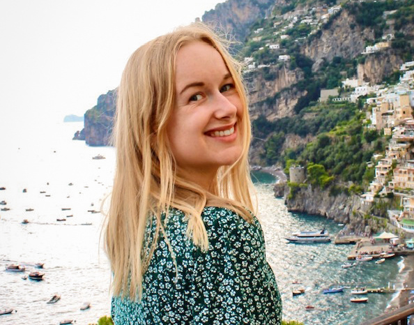 KI generiert: Eine Person lächelt vor einer Küstenlandschaft mit Booten und Häusern auf einem Hügel.