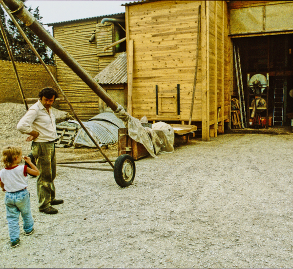 KI generiert: Ein Mann und ein Kind stehen neben einer landwirtschaftlichen Maschine vor einem Holzgebäude.