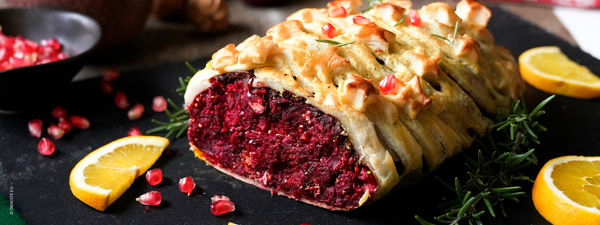 KI generiert: Das Bild zeigt ein gebackenes Gericht, das in Teig gehüllt ist und eine rote Füllung aufweist, vermutlich aus Rote Bete. Dekoriert ist es mit Rosmarin, Zitronenscheiben und Granatapfelkernen.