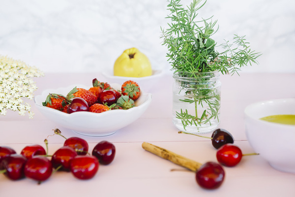 KI generiert: Frische Erdbeeren und Kirschen in einer Schale, mit Kräutern, Zitrone und Holunderblüten dekoriert.