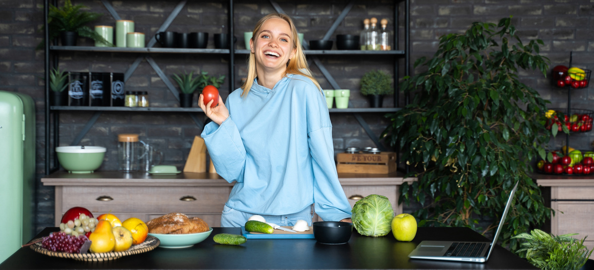KI generiert: Eine fröhliche Person steht in einer modernen Küche, umgeben von frischem Obst und Gemüse. Sie hält eine Tomate und vor ihr steht ein Laptop auf dem Tisch.