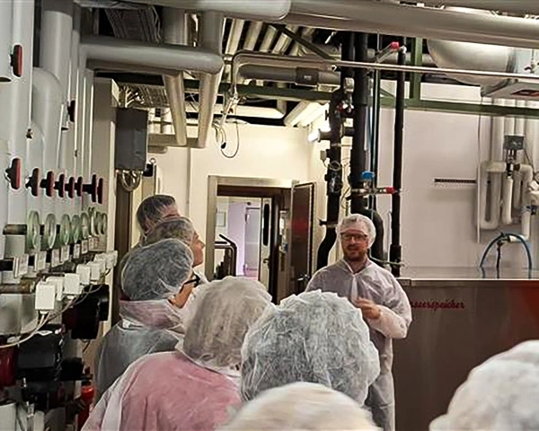 KI generiert: Menschen in Schutzkleidung bei einer Führung in einer Fabrik. Text: "Gemeinschaftsspeicher".