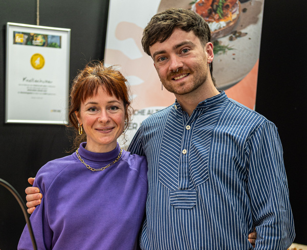 KI generiert: Zwei Personen posieren lächelnd vor einer schwarzen Wand mit einem Bild und einem gerahmten Zertifikat.