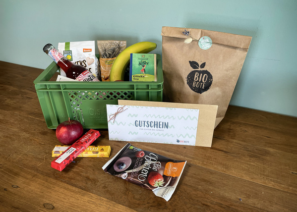 KI generiert: Eine grüne Kiste mit Bio-Produkten, einem Apfel, Schokolade und einem Gutschein. Text: "Gutschein für tollen Bio-Genuss".