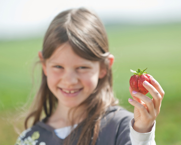 KI generiert: Ein Kind hält eine Erdbeere in der Hand und lächelt. Hintergrund ist verschwommen.