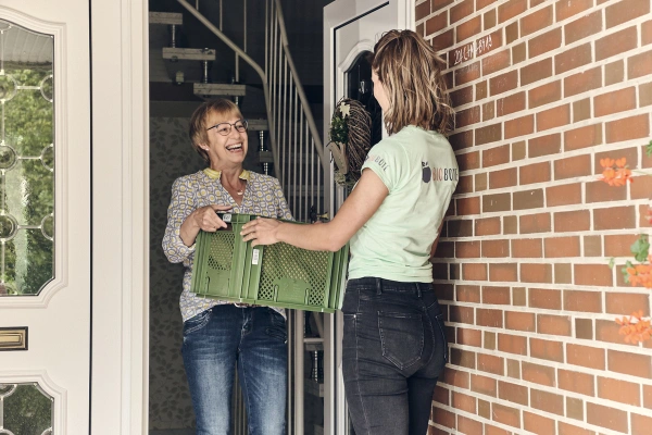 KI generiert: Zwei Frauen tauschen eine grüne Kiste an einer Haustür aus.
