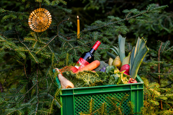 KI generiert: Grüne Kiste mit Gemüse, Obst und Wein in einem Nadelbaum mit Kerze und Deko.