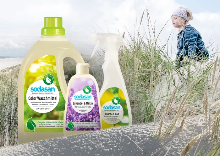 KI generiert: Sodasan Produkte vor sandiger Dünenlandschaft mit Kind. Text: "sodasan Color Waschmittel", "Lavendel & Minze", "Dusche & Bad".