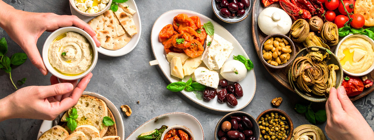 KI generiert: Mediterrane Leckereien wie Käse, Oliven, Hummus und Antipasti auf mehreren Tellern, zwei Hände greifen zu.