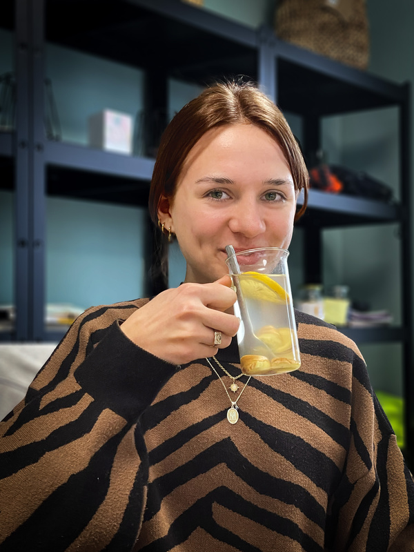 KI generiert: Eine Person hält ein Glas Wasser mit Zitronenscheiben und lächelt dabei in die Kamera. Im Hintergrund sind Regale zu sehen.