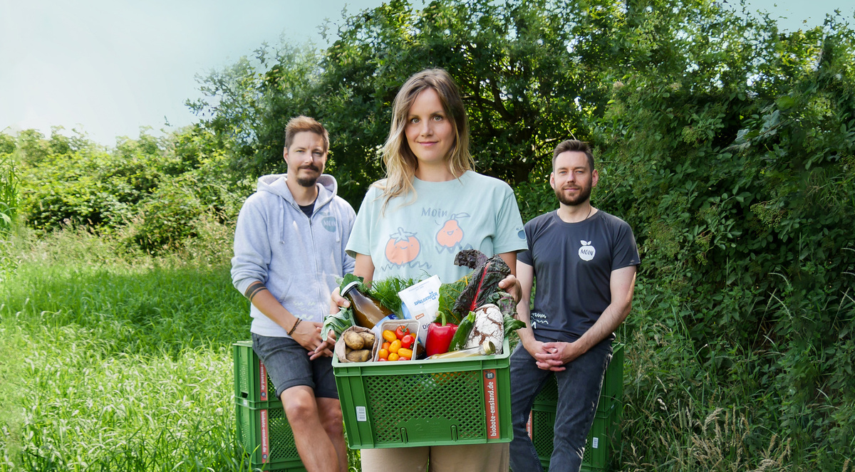 KI generiert: Drei Personen stehen draußen mit Kisten voller frischem Gemüse und Kräutern. Es gibt keinen Lesetext.