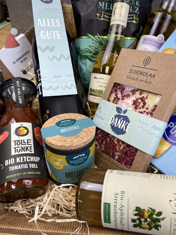 KI generiert: Das Bild zeigt einen Geschenkkorb mit verschiedenen Bio-Produkten, darunter Ketchup, Brotzeit-Salz, Öl und Schokolade. Auf den Verpackungen sind freundliche Botschaften wie "Alles Gute" und "Danke" zu sehen.