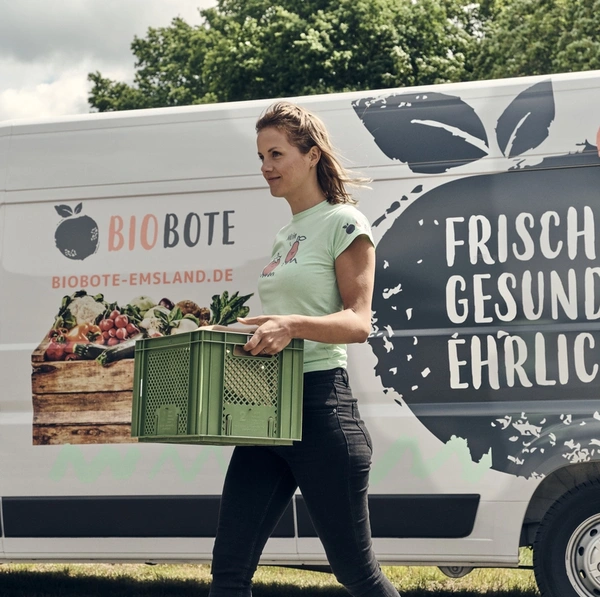 KI generiert: Eine Frau trägt eine Kiste vor einem Lieferwagen mit der Aufschrift "FRISCH GESUND EHRLICH".