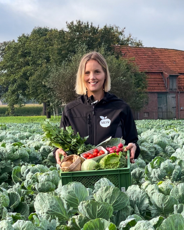 KI generiert: Eine Person steht in einem Gemüsefeld und hält eine Kiste mit frischem Gemüse. Im Hintergrund ist ein Bauernhof zu sehen.
