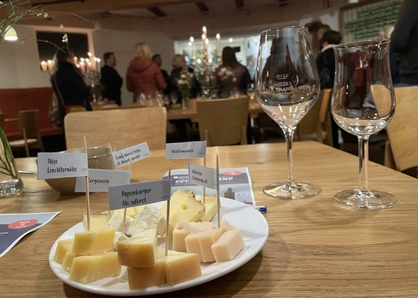 KI generiert: Käseplatte mit Beschriftungen auf einem Tisch, neben Weingläsern. Im Hintergrund Personen im Raum.