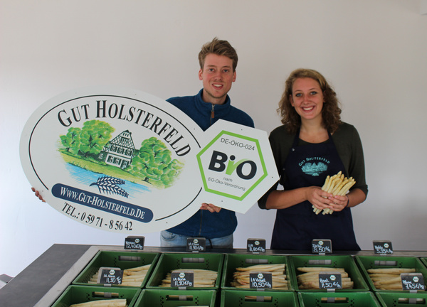 KI generiert: Zwei Personen präsentieren Spargel und ein "Gut Holsterfeld"-Schild mit Bio-Logo. Preise sind sichtbar.