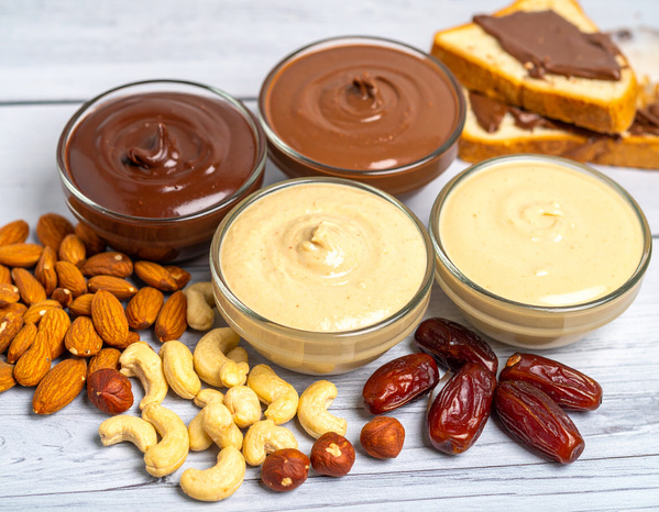 KI generiert: Verschiedene Nusscremes in Schalen mit Mandeln, Cashews, Datteln und Haselnüssen, Brotscheiben im Hintergrund.