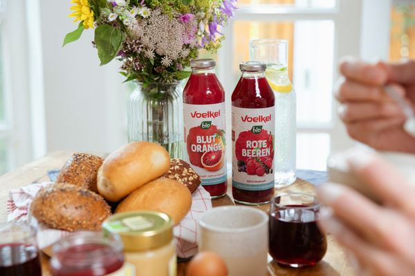 Frühstückstisch mit Voelkel-Säften, Blumen und Brötchen. Text: "Voelkel bio BLUTORANGE" und "ROTE BEEREN".